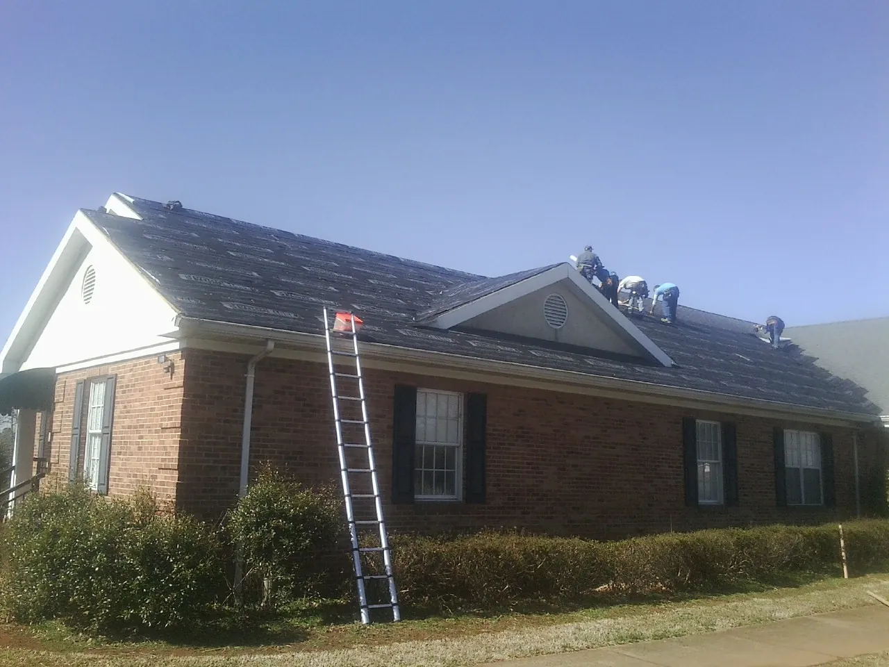 Crew tearing shingles off a brick ranch roof with a ladder set — Metal Roof Repair work by our crew in Mount Braddock