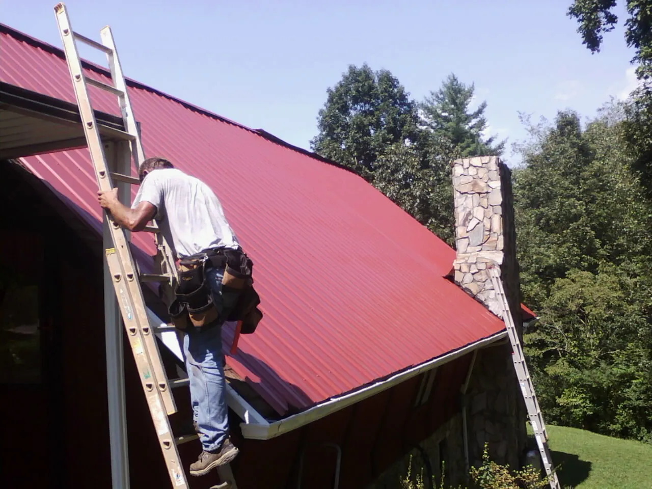 Roofer on a ladder beside a red metal roof and stone chimney — Soffit & Fascia Repair by Roofing Brothers in Mount Braddock